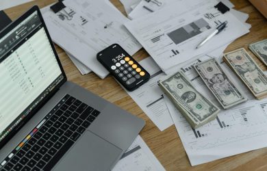 Laptop and smartphone with calculator on desk cluttered with financial documents and US dollar bills